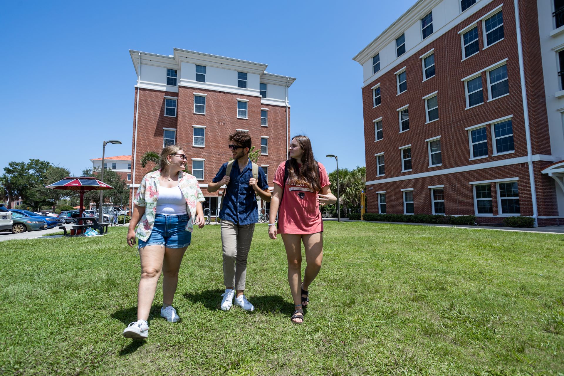 Three students walking and talking on a grassy area between two brick dormitory buildings on a sunny day
